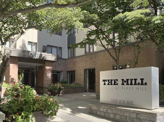 The Mill at First Hill is a modern building with a large entrance sign and is surrounded by greenery.