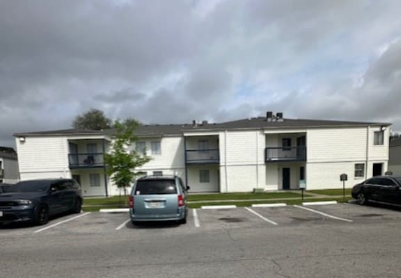 A parking lot in front of a white apartment building with a silver van and two black cars.
