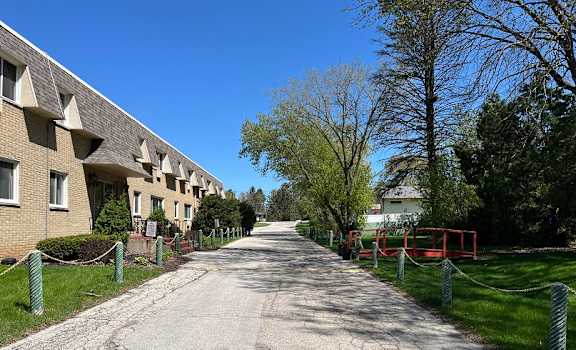A long, narrow road with a sidewalk on one side and a fence on the other, leading to a building.