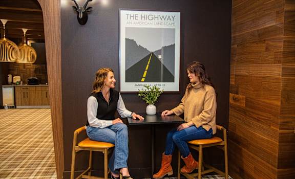 Two women sitting at a table in a room with a poster that says "The Highway" on it.