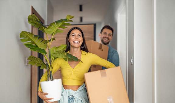 A woman is holding a potted plant and a cardboard box while a man is carrying another cardboard box.