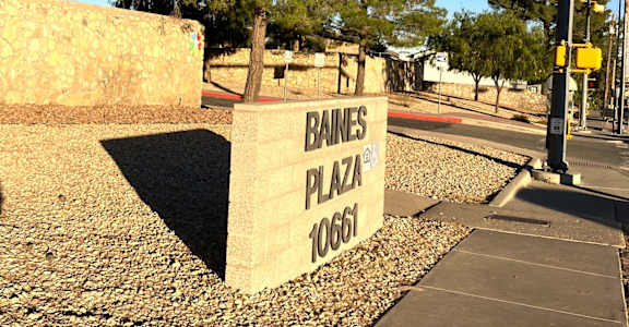 A stone sign that says Baines Plaza stands on a gravel area.