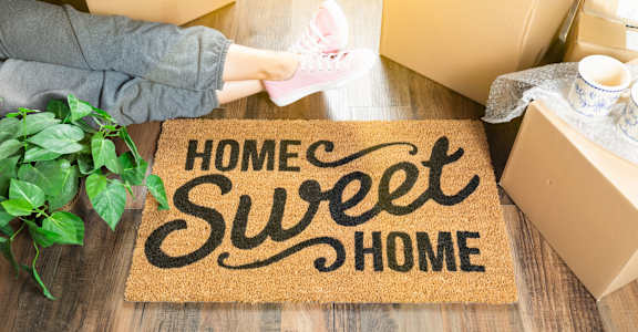 A person is unpacking boxes on a doormat that says "Home Sweet Home".