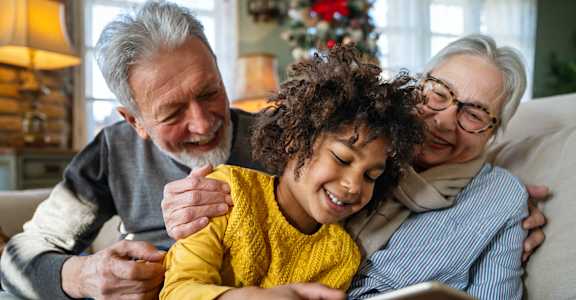 A family of three is sitting on a couch with a tablet in the middle person's hands.