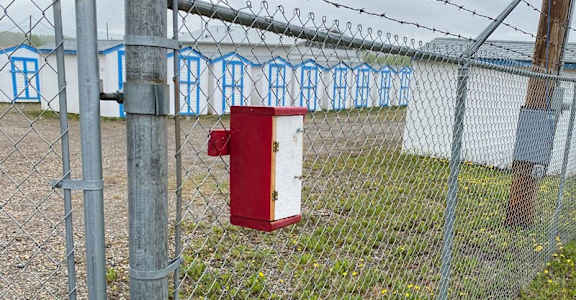 A red mailbox is attached to a chain link fence with barbed wire on top.