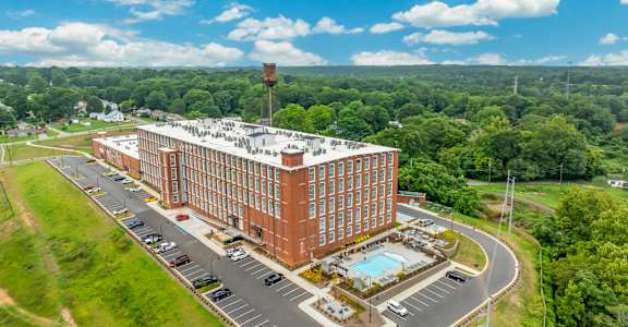 Loft Studios, Spartanburg, SC - Converse Mill Lofts - Aerial View Of A Large Brick Apartment Building With Parking Areas, Surrounding Greenery, and Nearby Homes.