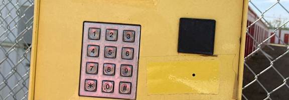A yellow box with a keypad and a small screen is attached to a chain link fence.