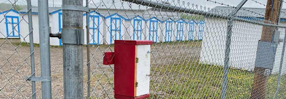 A red mailbox is attached to a chain link fence with barbed wire on top.