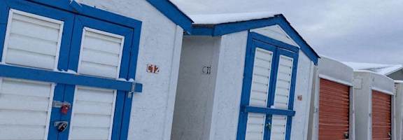 A series of storage units with blue doors are covered in snow.