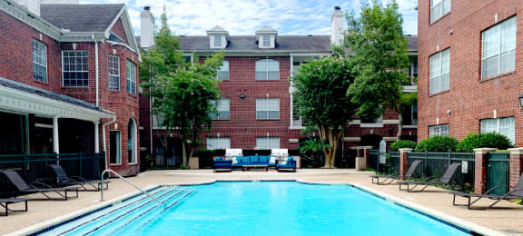Luxury apartments with red brick exteriors, large swimming pool with spacious sundeck, grill, beautiful landscaping, lush landscaping, chaste trees, and pool-side lounge seating at Tuscany Apartments in Houston.