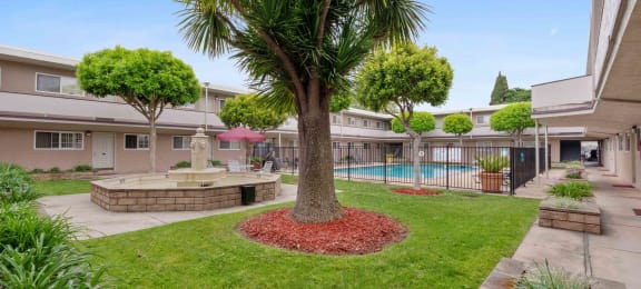 Apartments For Rent In San Lorenzo, CA - Lorenzo Commons - A Large Open Courtyard With Many Trees, A Fountain, And A Fenced In Pool Area