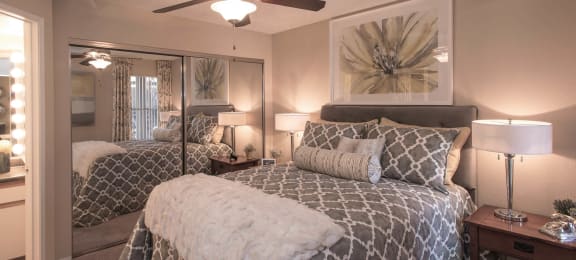 One and Two-Bedroom Apartments in Ahwatukee, AZ - Pacific Bay Club - Bedroom with Mirrored Closet Doors, Carpet Floors, Ceiling Fan, Cozy Bed, Stylish Decor, and Door to Bathroom with Wood-Style Plank