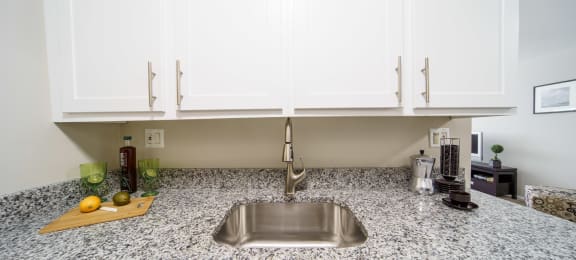 Stainless steel sink with white upper cabinets and  grey and white stone counters.