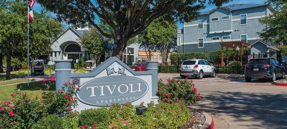 Apartments in North Dallas - Tivoli - Community Entrance with Gray Building, Entrance Sign, Lush Greenery, and Parking Lot