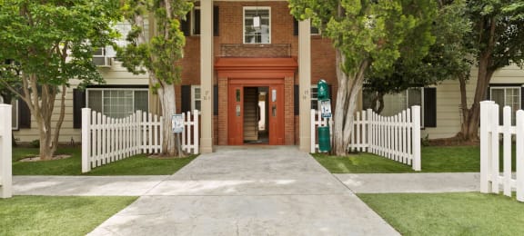 Apartments for Rent in Van Nuys CA - Colonial Manor - Exterior View Of Apartment Complex Entrance Featuring Stone Walkway, Lush Green Grass and Trees, And White Picket Fence