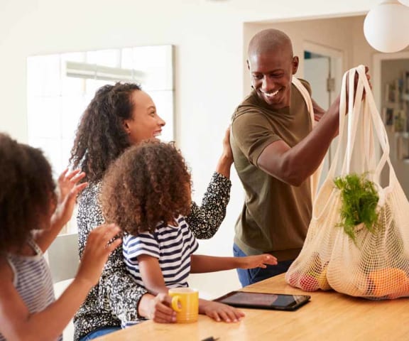 a family of four is gathered around a kitchen table with a grocery bag of vegetables