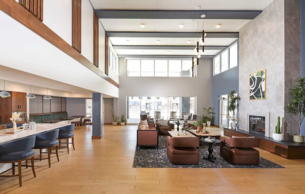 Shared clubhouse lounge with tall windows and seating — view 2 at Juniper Square 55+ Apartments.