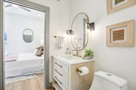 A bathroom with a toilet, sink, mirror, and a bedroom in the background.