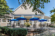 A building with a beige roof and white columns is surrounded by a black fence and has blue umbrellas on the patio.