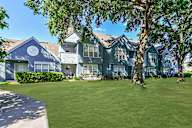 An apartment building with blue siding and white trim, in front of well manicured green grass and mature trees.