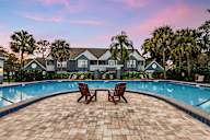 Two modern pool chairs are placed on a brick paver patio in front of a swimming pool, with an apartment building in the background..