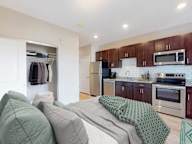 A bedroom with a bed covered in a green blanket and a kitchen area with a refrigerator, microwave, and oven.