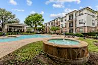 A large outdoor swimming pool with a hot tub in the middle of a grassy area in front of apartment buildings.