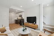 Modern open-plan living space with hardwood flooring, featuring a cozy sitting area with a white round marble-top coffee table, sleek TV stand, and an understairs white door.
