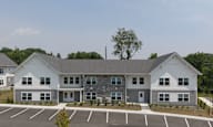 A large white building with a grey roof and a parking lot in front.