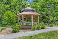 A wooden gazebo with a black roof is surrounded by greenery and a lamp post.