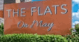 A sign that says "The Flats One Way" in front of a hedge.