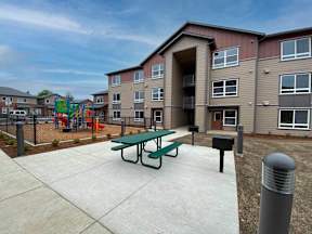A playground with a slide and swings is in front of a building with a green picnic table in front of it.