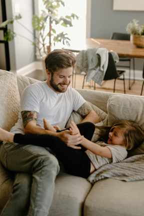 A man and a little girl are sitting on a couch, the man is holding a phone.