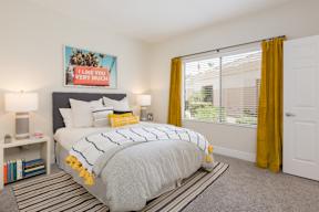 A bedroom with a bed, a nightstand, a lamp, a picture frame, a bookshelf, a rug, and a window with yellow curtains.