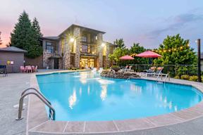 A swimming pool in front of a house with a stone wall and a patio with chairs and umbrellas.