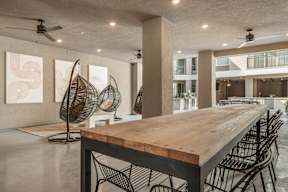 A long wooden table with chairs is in the foreground of a spacious room with a fan and artwork on the wall.