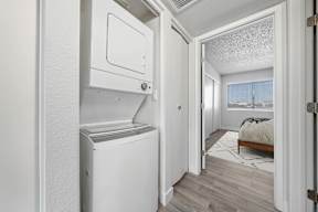 A white washing machine is in a small room with a bed in the background.