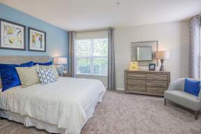 A bedroom with a white bed, a dresser with a mirror, and a chair with a blue pillow.