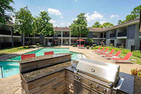 A grill is in the foreground of a photo showing a pool and apartment complex.