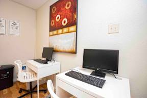 A white desk with a black monitor and keyboard sits in a room with a painting on the wall.