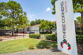 A sign for Three Corners North stands in front of a building and trees.