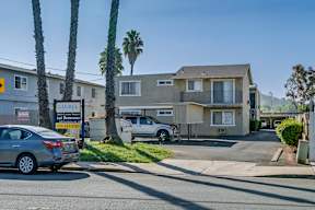 A street view of a residential area with houses, cars, and palm trees.