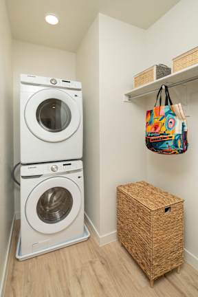 A small laundry room with a double washer and dryer, a wicker basket and a bag hanging on the wall.