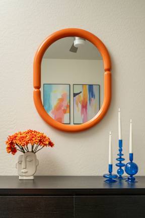A round orange mirror with a painting behind it and a vase with orange flowers in front of it.