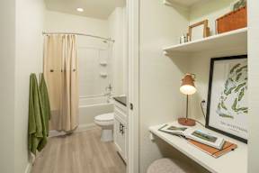 A bathroom with a toilet, a shower with a curtain, a shelf with a lamp and a picture frame.