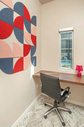 A red, blue and grey abstract art piece hangs on a wall above a desk with a black chair.