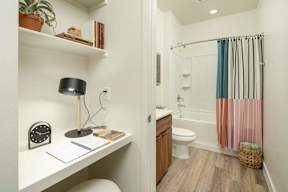 A bathroom with a toilet, a sink, a shelf, a clock, a potted plant, a lamp, a towel rack, a basket, and a shower curtain.