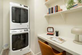 A white microwave oven is placed next to a white desk with a chair and a clock on it.