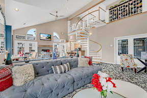 A grey tufted sofa with a red throw pillow sits in a living room with a spiral staircase.