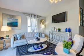 A living room with a white couch, a coffee table, and a flat screen TV mounted on the wall.